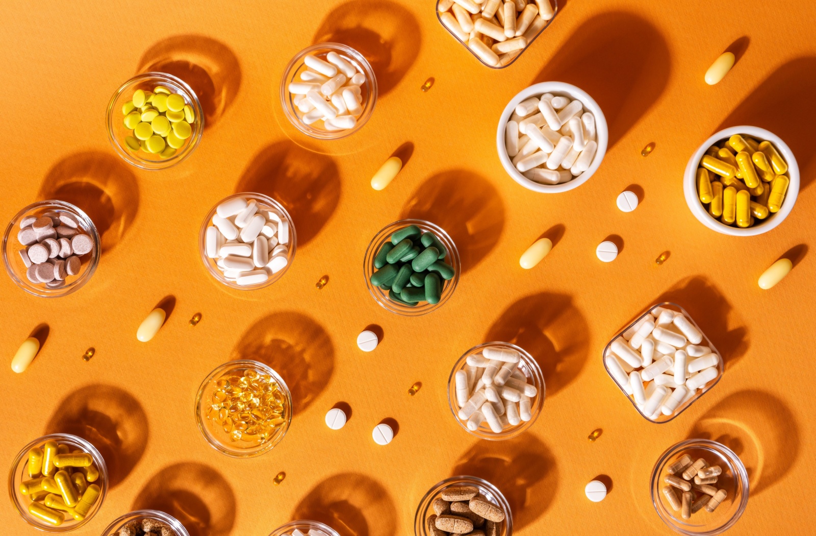 Various multivitamin capsules and tablets in glass bowls arranged on an orange background with shadows