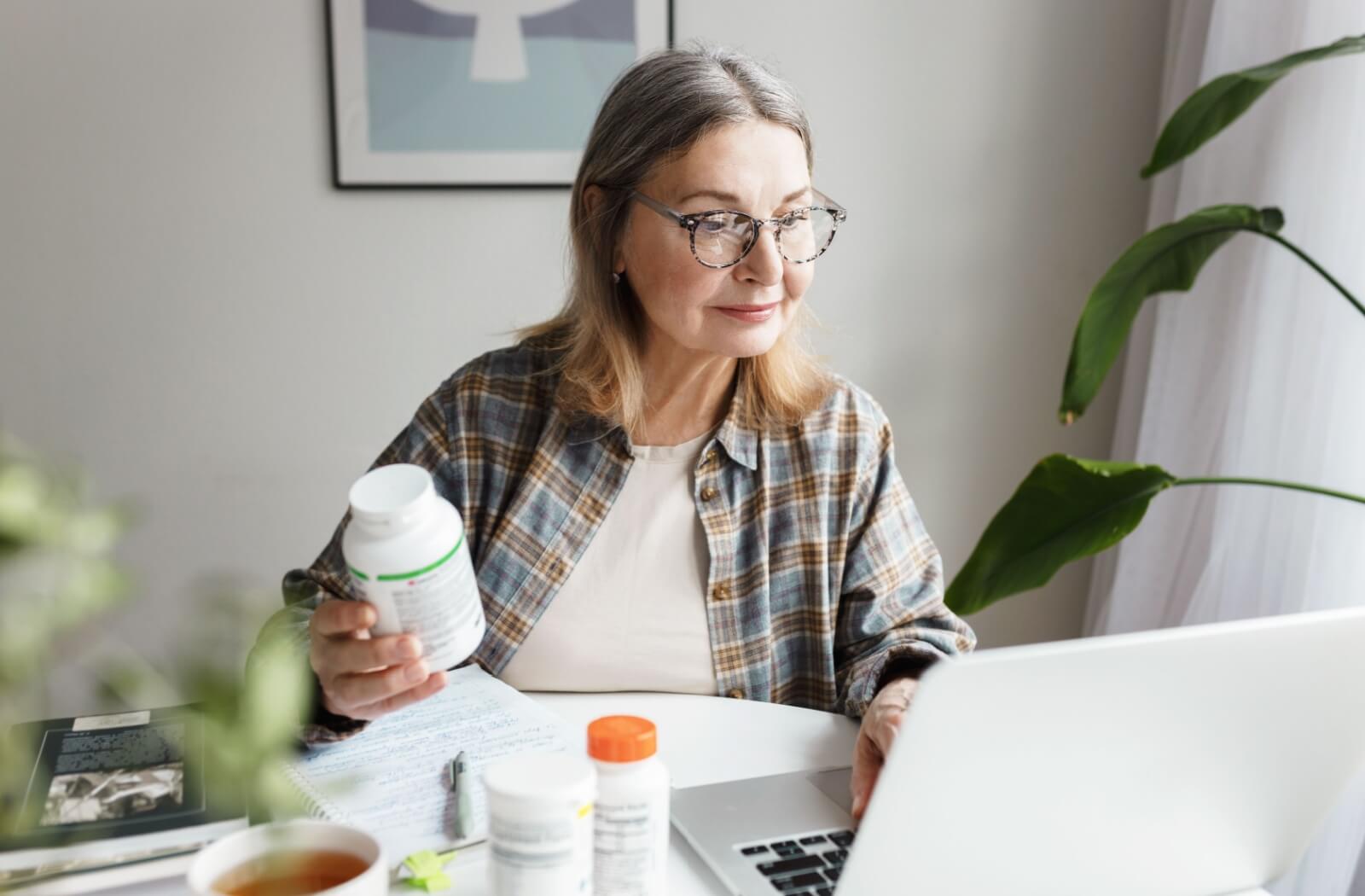 Older adult researching vitamins online while holding a supplement bottle at a desk with a laptop and notes