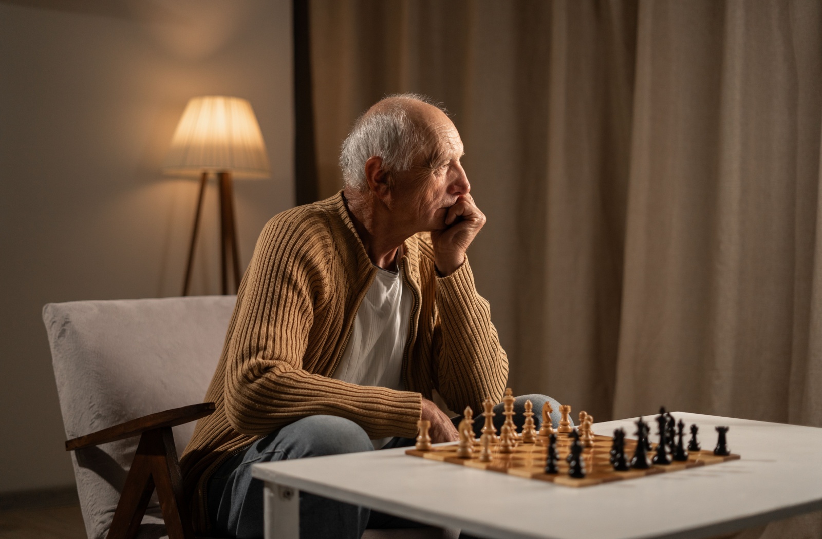 A senior with dementia in a brown cardigan sits alone at home in front of a chessboard with no one to play against