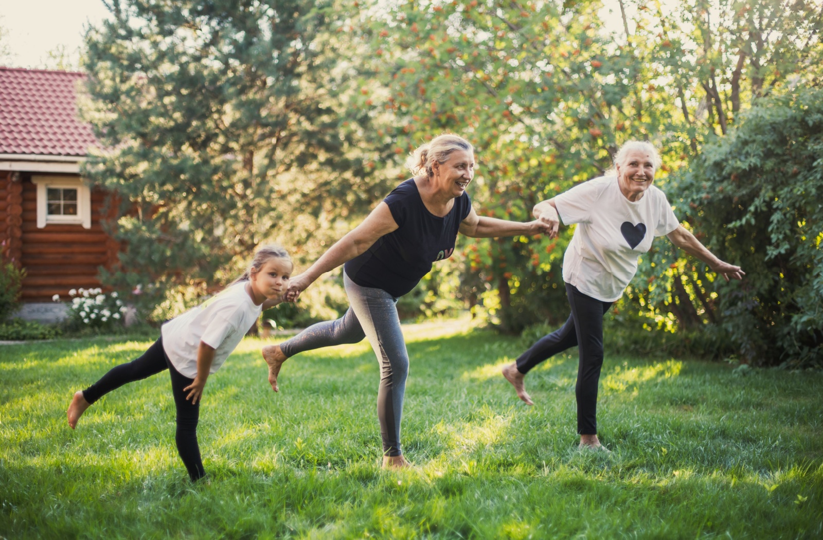 Three family members, each from a different generation, hold hands while balancing barefoot on one leg in the backyard