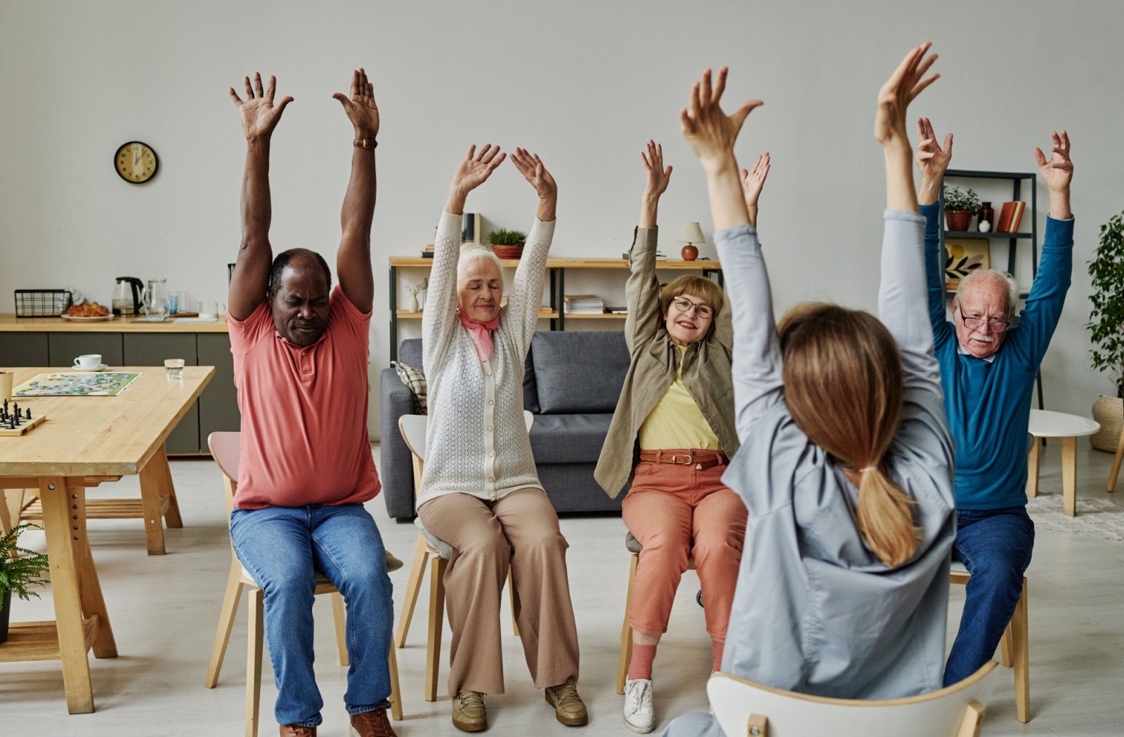 A group of seniors sit in chairs and stretch their arms up, under the guidance of a yoga instructor at a senior living community.