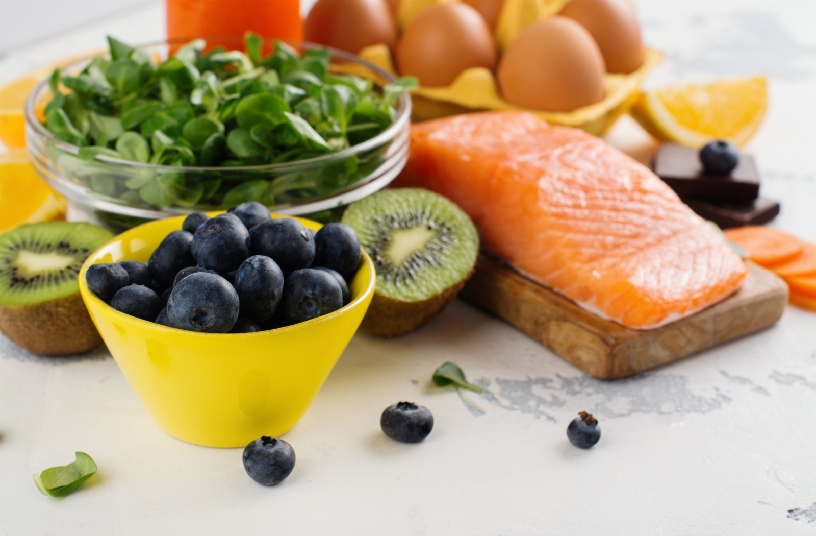 A selection of healthy foods, including blueberries, a kiwi, spinach, salmon, eggs, and orange slices.