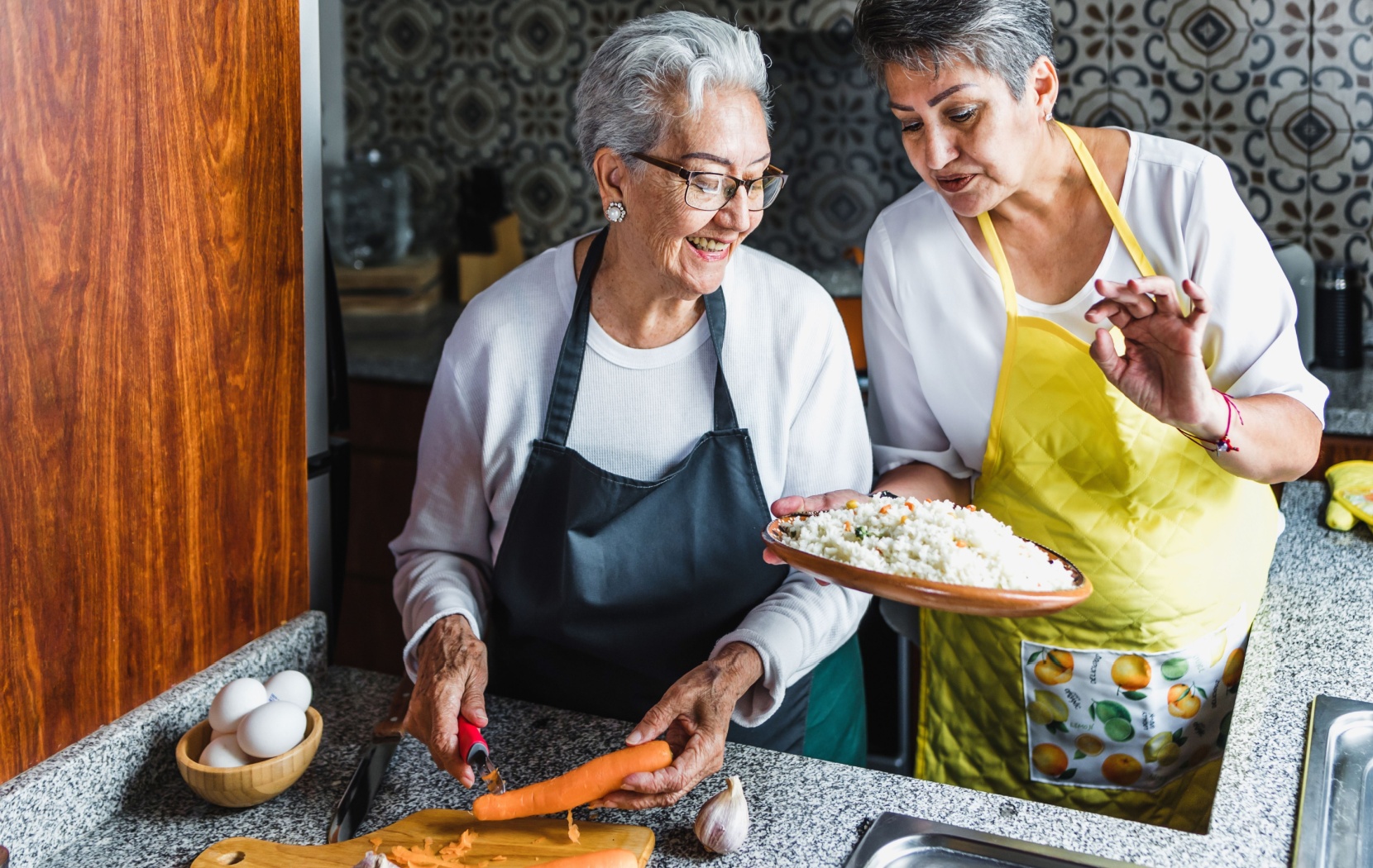 A senior holds a plate of cooked rice, as another senior peels carrots.