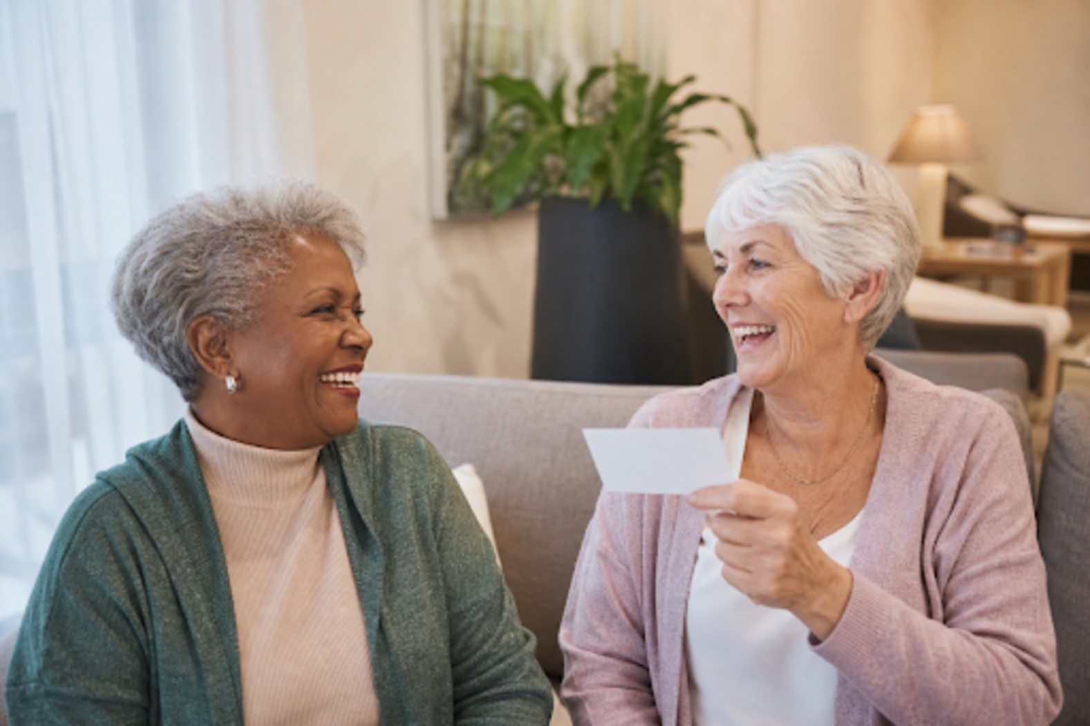 Two older adults sit on a couch laughing together, one holding a small photo or card.
